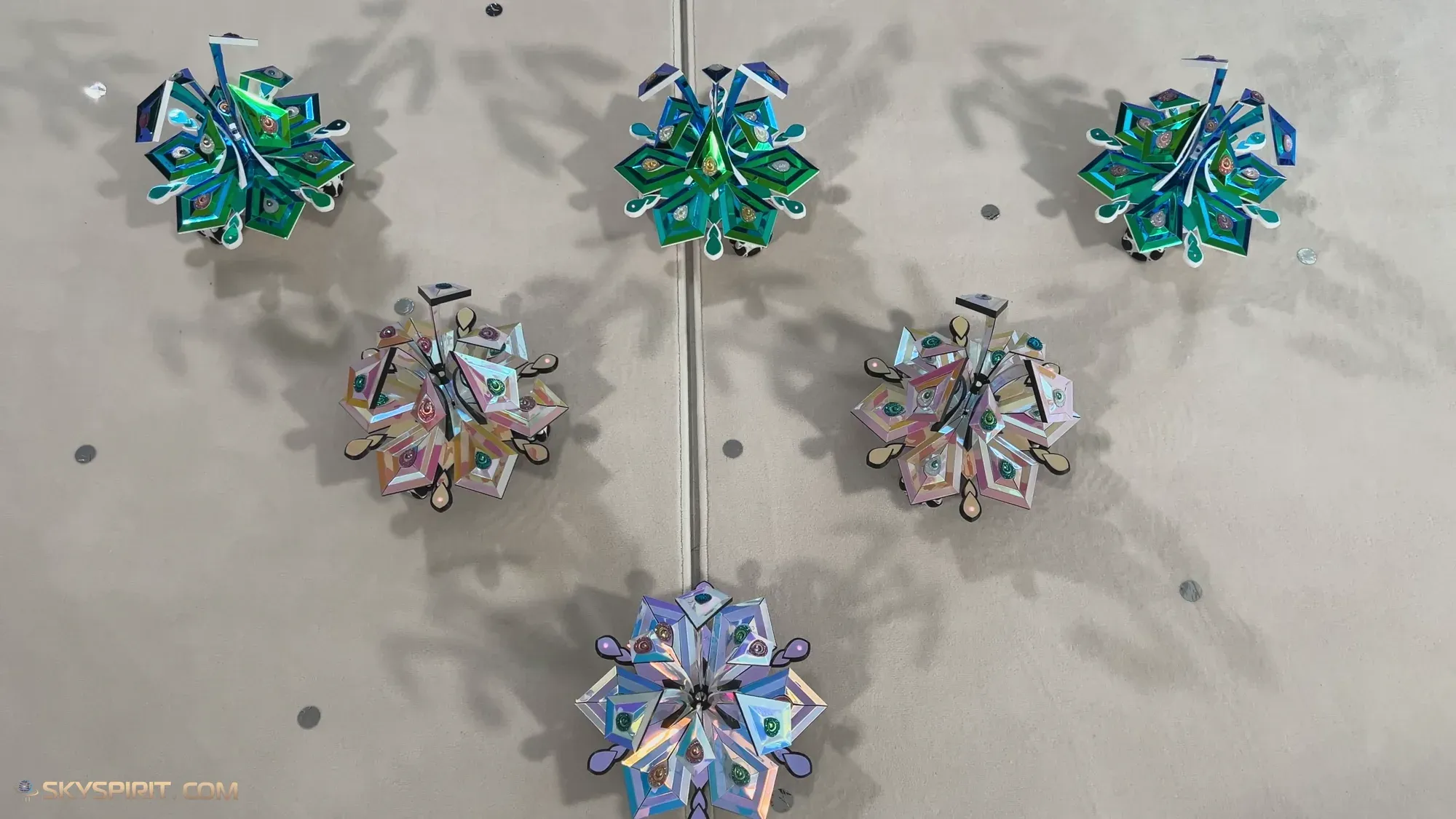 Six iridescent star-flower robots in green, blue, pink and gold, on omni-wheels and in formation on a pale grey floor, seen from above