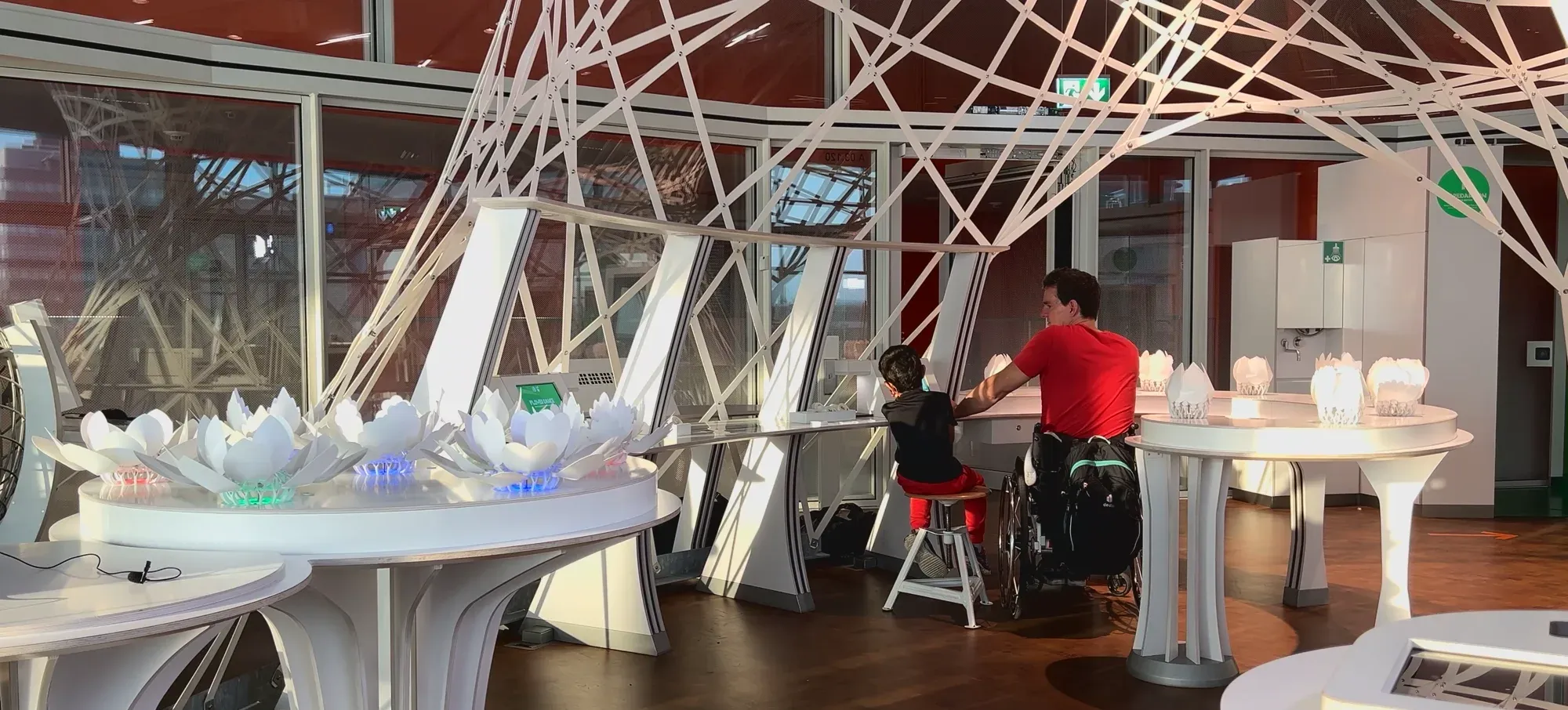 A visitor in a wheelchair working at a station next to a child on a stool, blossoms lit along the white benches