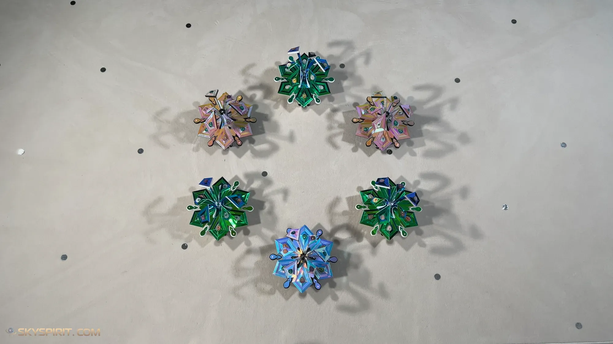 Six star-flower robots in green, blue, pink and gold forming a circle, seen from above