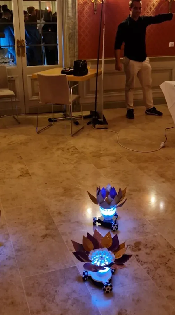 The man raising his arm at the back of the hall while two flower robots light up blue on the floor