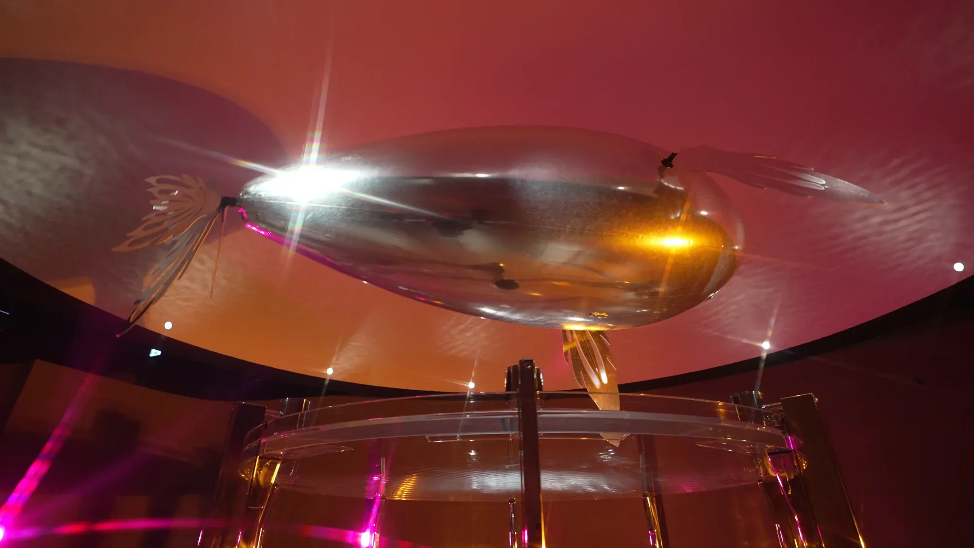 A SkyVoyager seen from below, hovering over a glass drum under a warm orange ceiling
