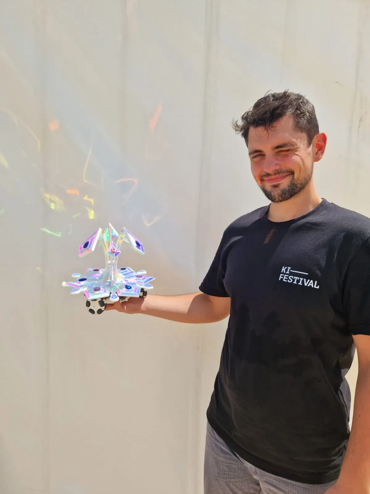 A man in a KI Festival T-shirt holds one of the small iridescent blossom robots on his open palm